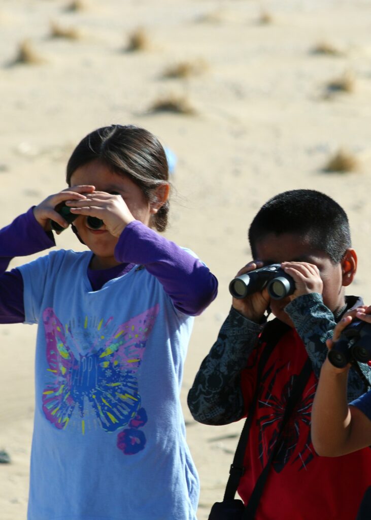 Two second graders looking through binoculars at birds on a beach